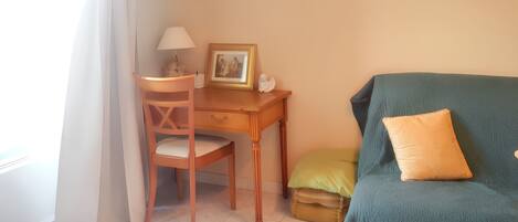 A room with a wooden desk, a chair, a framed picture, a lamp, and a green sofa with a cushion.