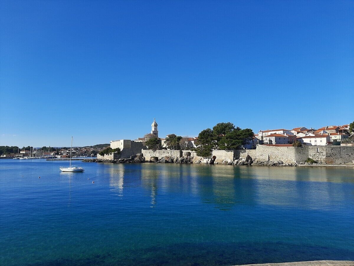 Un pueblo costero con mar azul cristalino, un edificio histórico con torre y un pequeño velero.