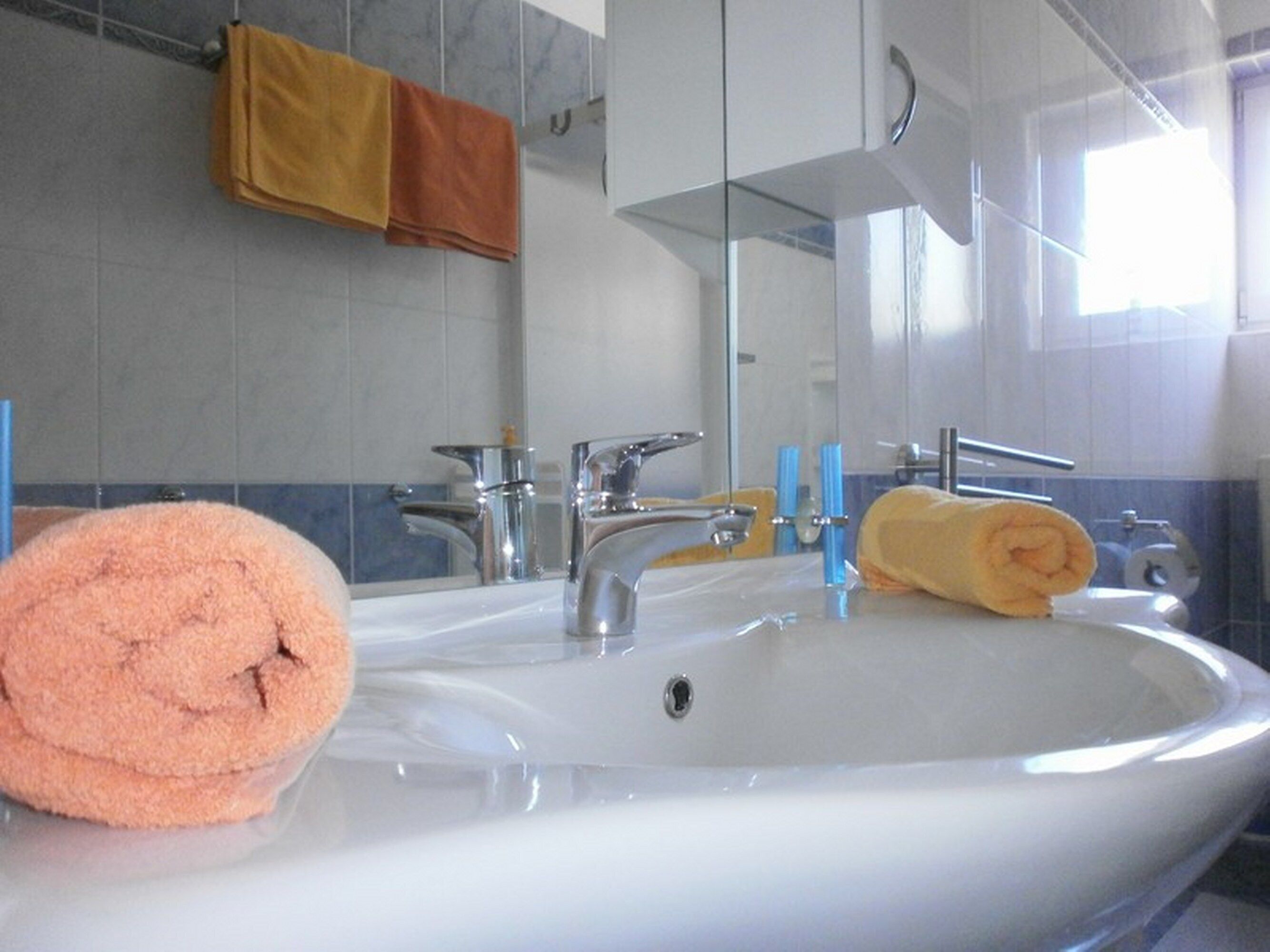 Ein Badezimmer mit einem weißen Waschbecken, zwei Wasserhähnen, einem Spiegel und Handtüchern an der Wand.