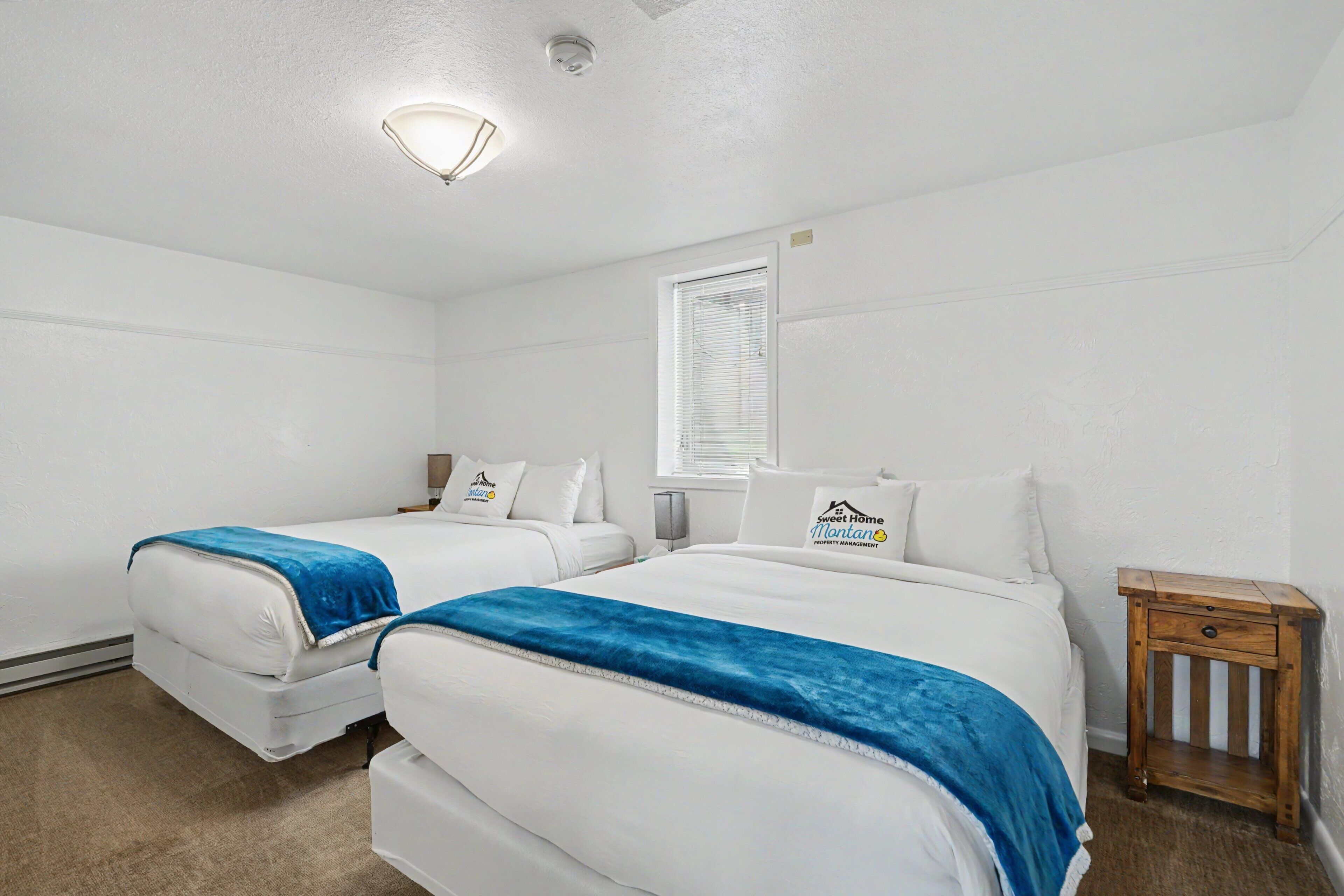 A hotel room with two beds, a small wooden nightstand, a window with blinds, and a ceiling light.