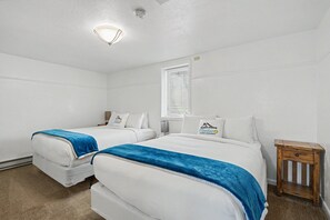 A hotel room with two beds, a small wooden nightstand, a window with blinds, and a ceiling light. - Huge 3-bedroom home just 1 minute from Yellowstone. Legacy West (West Yellowstone)