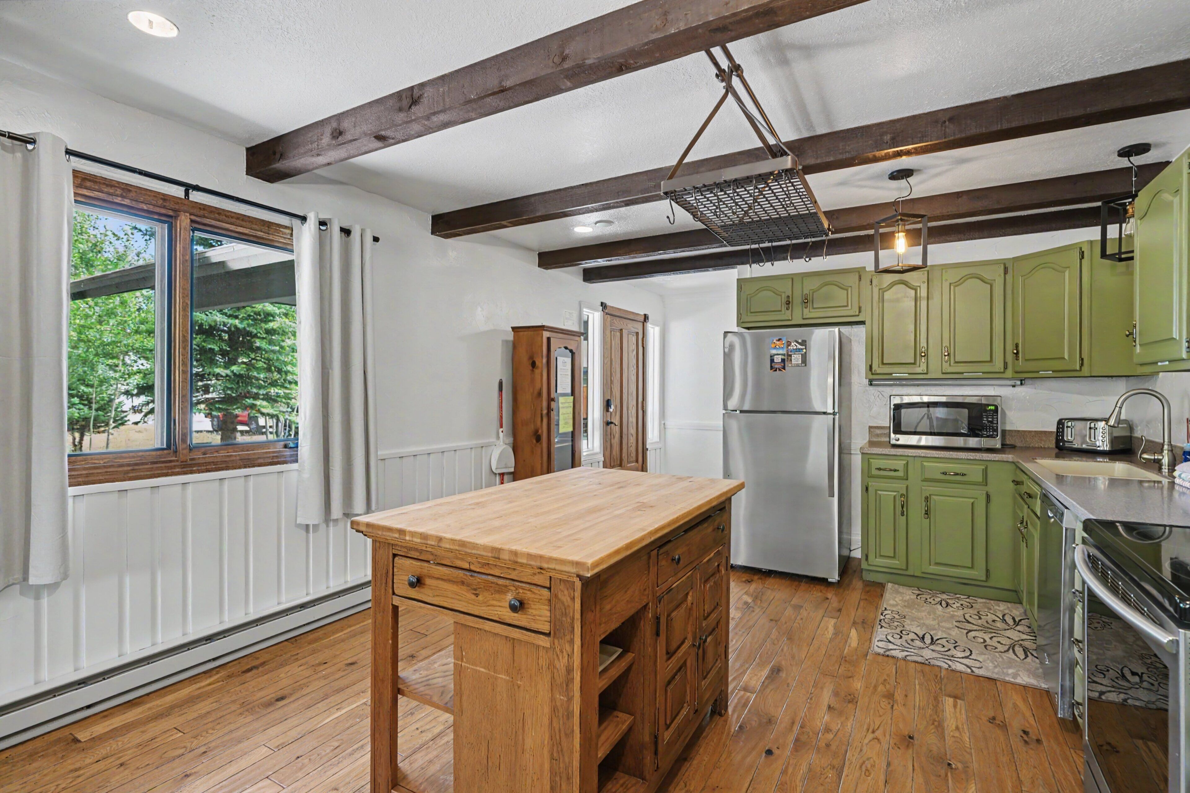 A kitchen with wooden flooring, green cabinets, a wooden island with drawers, a refrigerator, a microwave, and a hanging light fixture.