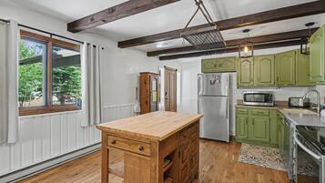 A kitchen with wooden flooring, green cabinets, a wooden island with drawers, a refrigerator, a microwave, and a hanging light fixture.