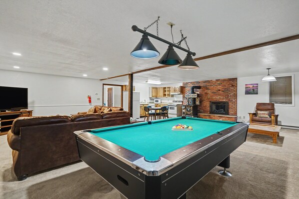 Une table de billard dans un salon, avec un canapé en cuir marron et un téléviseur en arrière-plan.