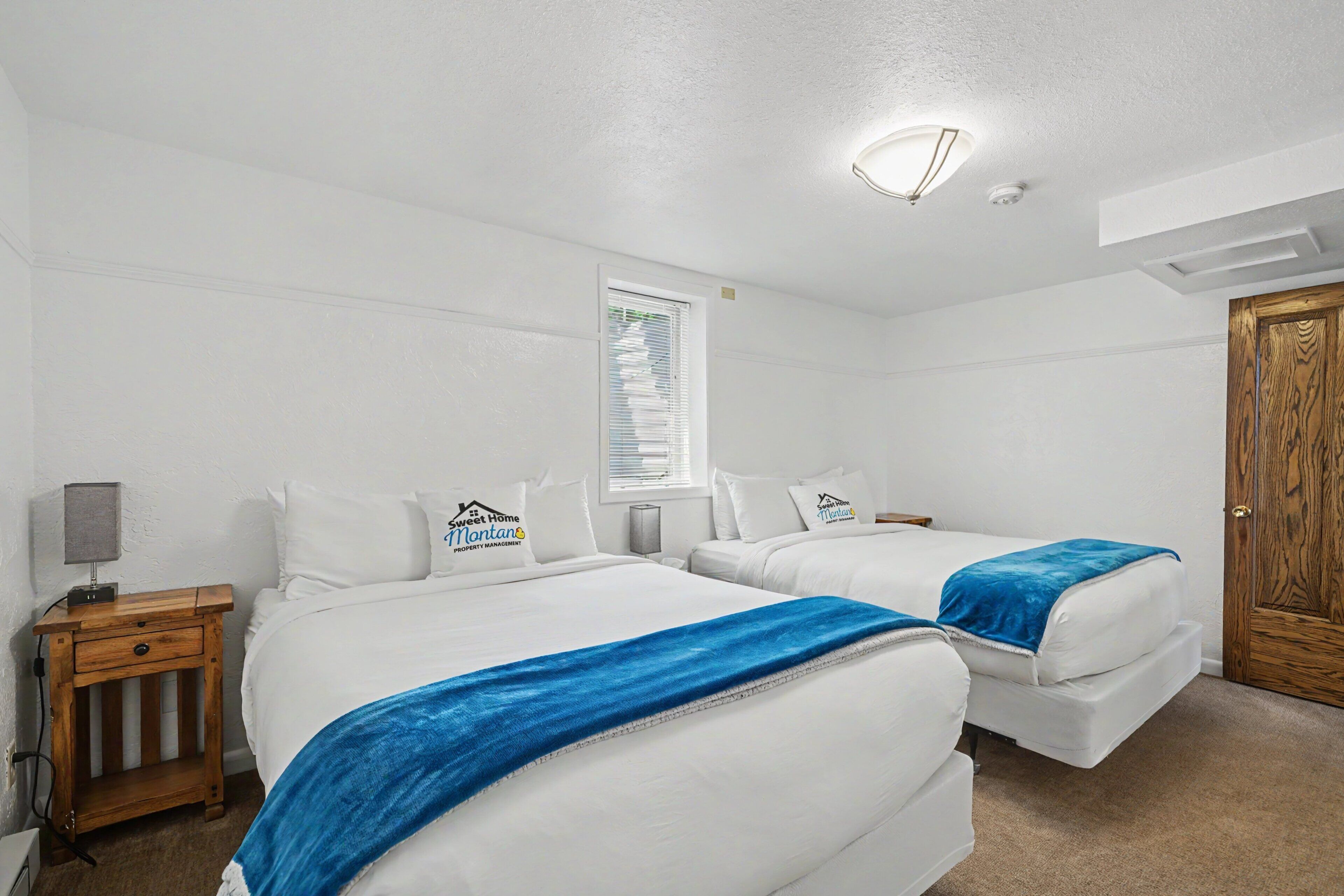 A hotel room with two beds, a desk, a lamp, and a window with blinds.