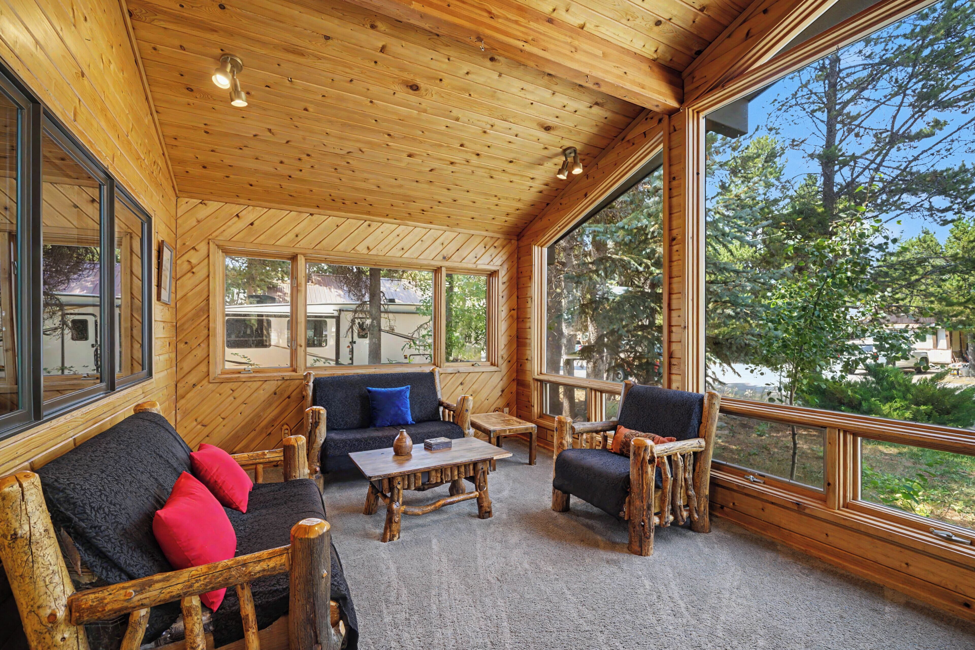 Un interior de madera con sofá, sillas y una mesa, ventanas amplias y vistas a árboles y un vehículo afuera.