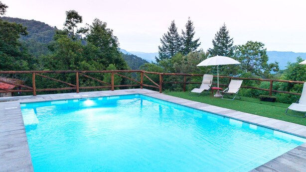 Ein rechteckiger Pool mit Holzterrasse und Metallgeländer. Zwei weiße Sonnenschirme und zwei Liegestühle sind vorhanden.