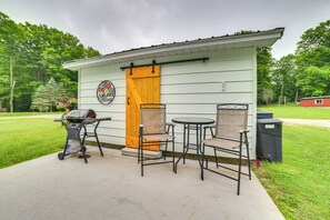 A small building with a wooden door, two chairs, a table, and a grill outside. - Visit Fife Lake & Traverse Bay! Farmhouse Hideaway (Fife Lake)
