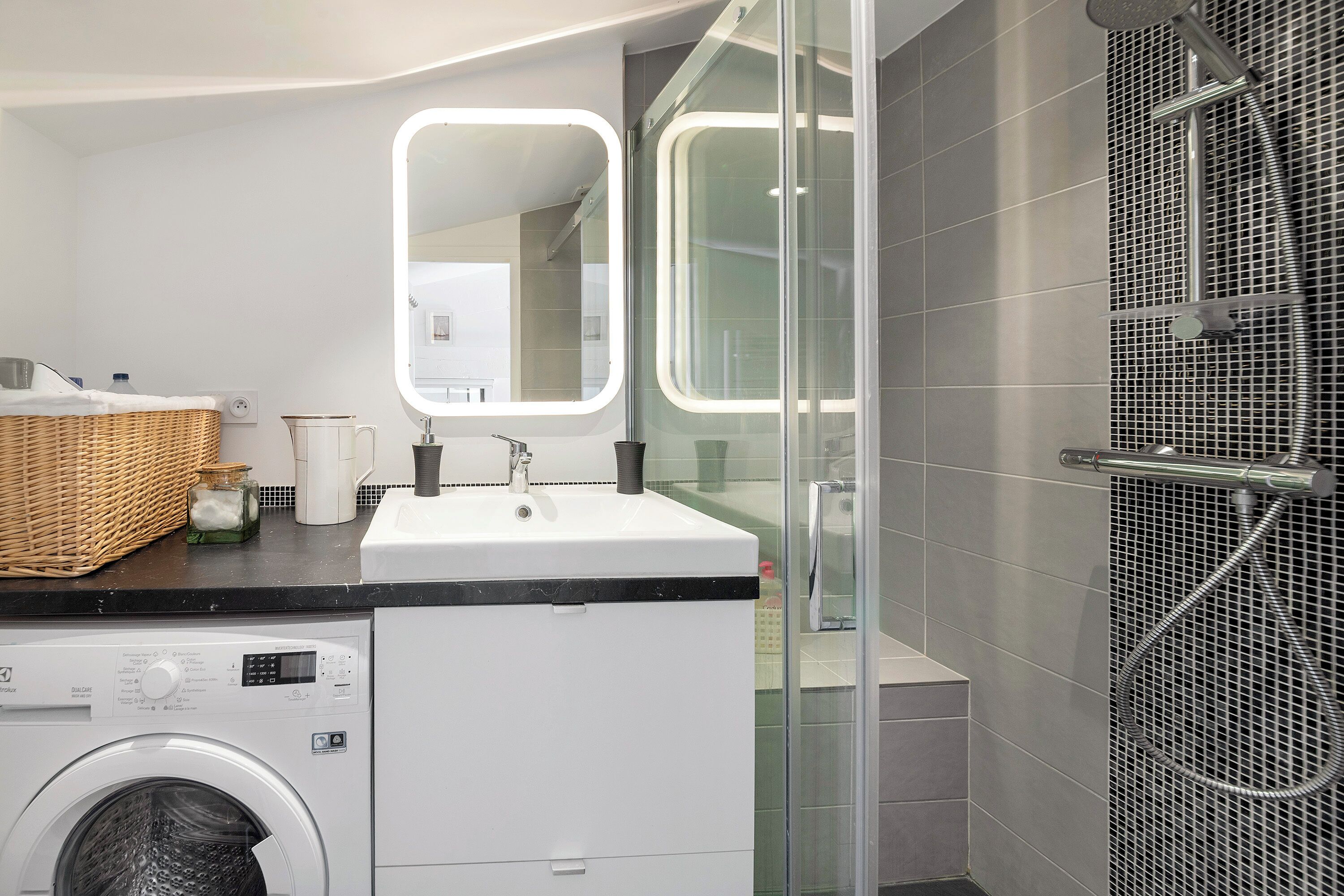Une salle de bain moderne équipée d’un lave-linge, d’un lavabo avec miroir et d’une cabine de douche.