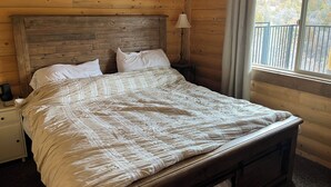 Un dormitorio de cabaña de madera con una cama, almohadas, una mesita de noche, una lámpara y una ventana con cortinas.