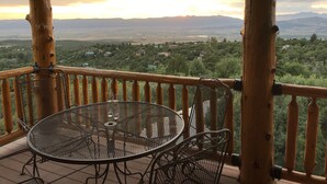 A wooden deck with a metal table and chairs, overlooking a scenic landscape with mountains and scattered houses.