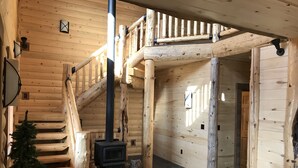 Interior de una cabaña de madera con una escalera central, una chimenea y un pequeño árbol.