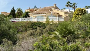 Una villa de estilo mediterráneo con techo de tejas, barandillas de balcón blancas y exuberante vegetación alrededor de la propiedad.