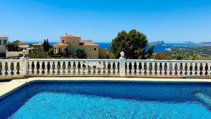 Una piscina con balaustrada blanca, vista a un pueblo costero y cielo azul despejado.