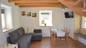 Una sala de estar acogedora con un sofá gris, una mesita pequeña, una mesa de comedor de madera con sillas, una ventana con persianas y un televisor montado.