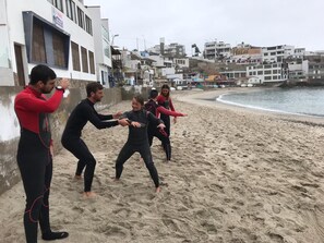 A group of people in wetsuits on a sandy beach with buildings in the background.