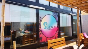 A modern building with a large glass facade, a wooden pergola, and outdoor seating. - Surfari Punta Rocas (Punta Negra)
