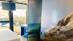 A room with a bed, a TV, a view of a beach, and a large painting of a sailboat. - Surfari Punta Rocas (Punta Negra)