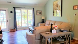 Un salon avec un canapé beige, une petite table avec une lampe, un tableau encadré au mur et une fenêtre avec des rideaux.
