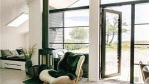 A room with a large sliding glass door leading to an outdoor area, a cushioned chair with a fur throw, a small seating area with cushions, and a potted plant.
