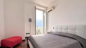 A bedroom with a bed, a red bench, a bedside table, and a view of the outdoors. - Larius Apartment Carate Urio (Carate Urio)
