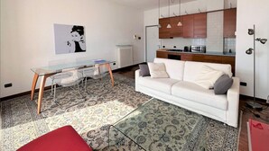 A modern living room with a white sofa, a glass dining table, and a red ottoman. There is a kitchen area with wooden cabinets and a microwave. - Larius Apartment Carate Urio (Carate Urio)