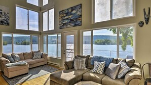 A spacious living room with a large sectional sofa, a coffee table, and a view of a lake through large windows. - Waterfront DeRuyter Home w/ Private Dock! (DeRuyter)