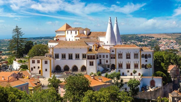 Un edificio histórico con múltiples fachadas blancas, una imponente torre campanario y un patio con una fuente.