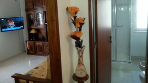 Una sala de estar con televisor, armarios de madera, un jarrón con flores y una puerta que da a un baño.