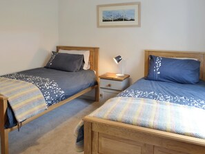 Une chambre à coucher avec deux lits en bois, chacun muni d’un oreiller et d’une couverture. Une table de chevet avec une lampe se trouve entre les deux lits. Un tableau encadré est accroché au mur, au-dessus des lits.