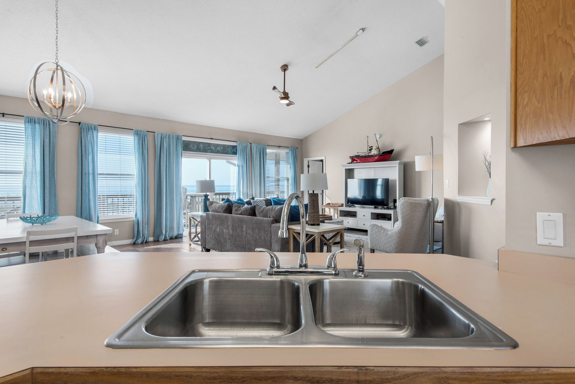 A modern kitchen with a double sink, a dining area with a table and chairs, a TV, and a view of the ocean.