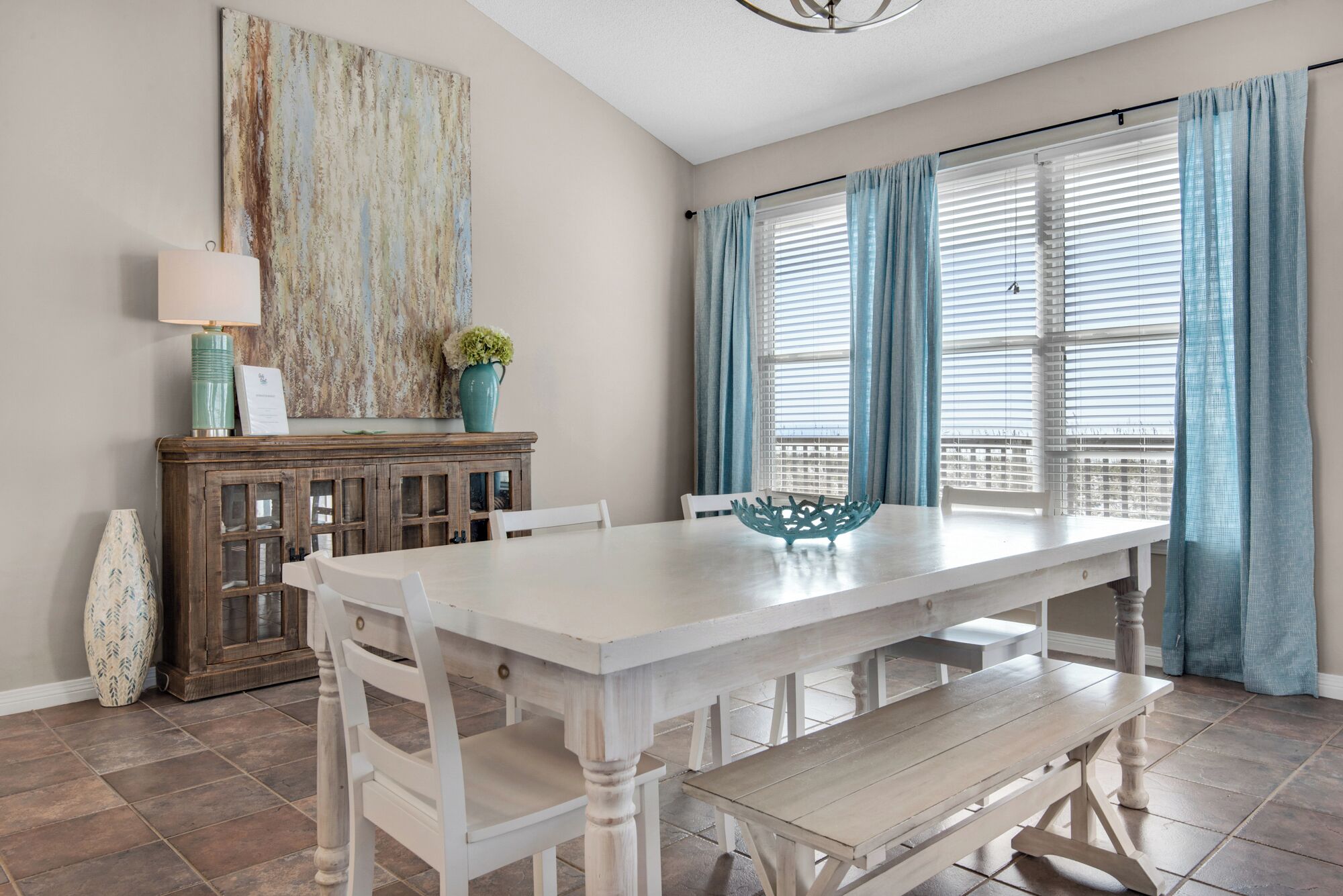A dining area with a white table, benches, and a decorative bowl. There is a cabinet with a vase and a lamp on top.