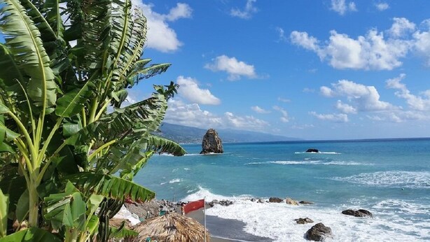 A beach with clear blue water, a rocky outcrop, and lush green foliage. - Villa Anna – Sea view attic with terrace, just minutes from the beach (Fuscaldo Marina)