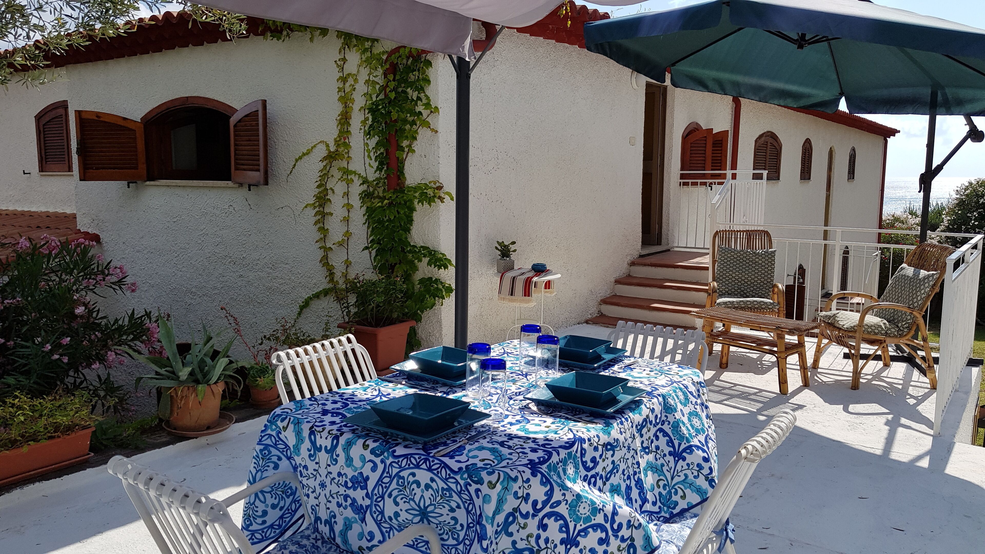 Un patio con una mesa redonda dispuesta para comer, rodeado de sillas blancas y un mantel azul y blanco estampado.