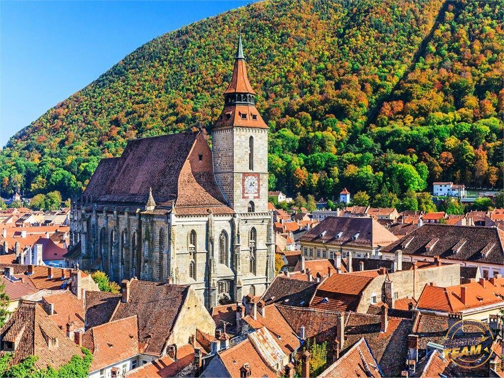 A historic church with a prominent tower and a clock, surrounded by traditional buildings and a hillside in the background.