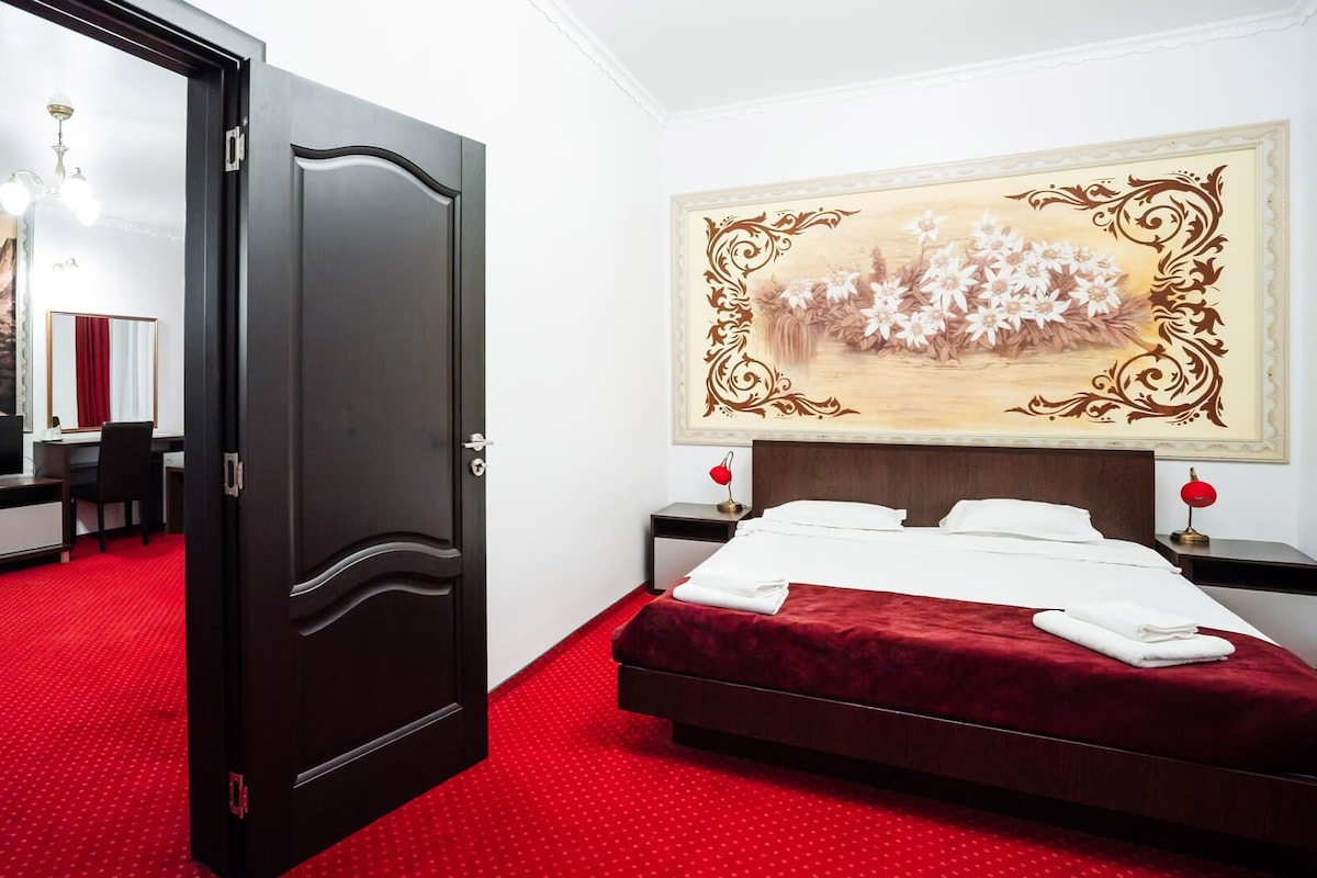 A hotel room with a red carpet, a bed with white linens, a large floral painting on the wall, a desk with a computer, and a television.