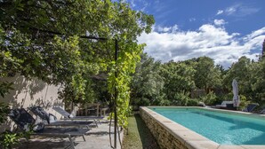 A swimming pool with lounge chairs and a pergola, surrounded by trees and a stone wall.