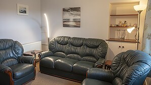 Ein Wohnzimmer mit einem schwarzen Ledersofa, einem passenden Sessel und einem kleinen Tisch. An der Wand hängt ein gerahmtes Bild und auf einem Beistelltisch steht eine Lampe.