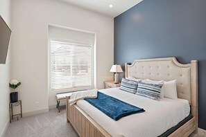A bedroom with a large bed, a bedside table with a lamp, a window with blinds, and a small plant on a side table.