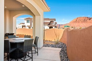 An outdoor patio with a dining table and chairs, a grill, and a view of neighboring buildings and a mountain.