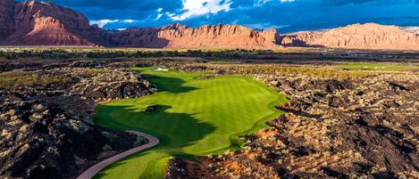 A golf course with a winding path, surrounded by rocky terrain and mountains.