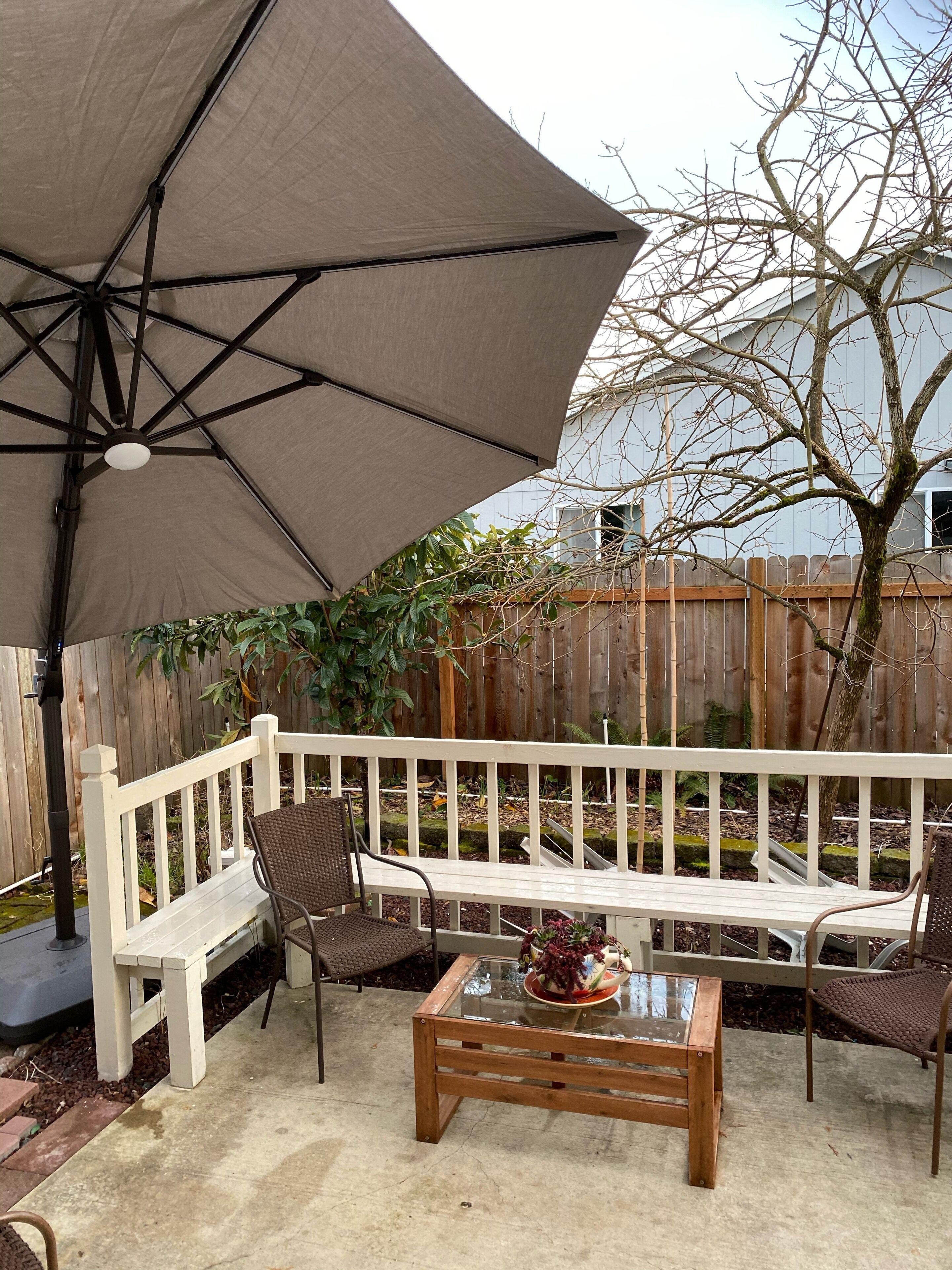 An outdoor patio with a white wooden bench, two wicker chairs, a glass-top coffee table, and a large umbrella providing shade.