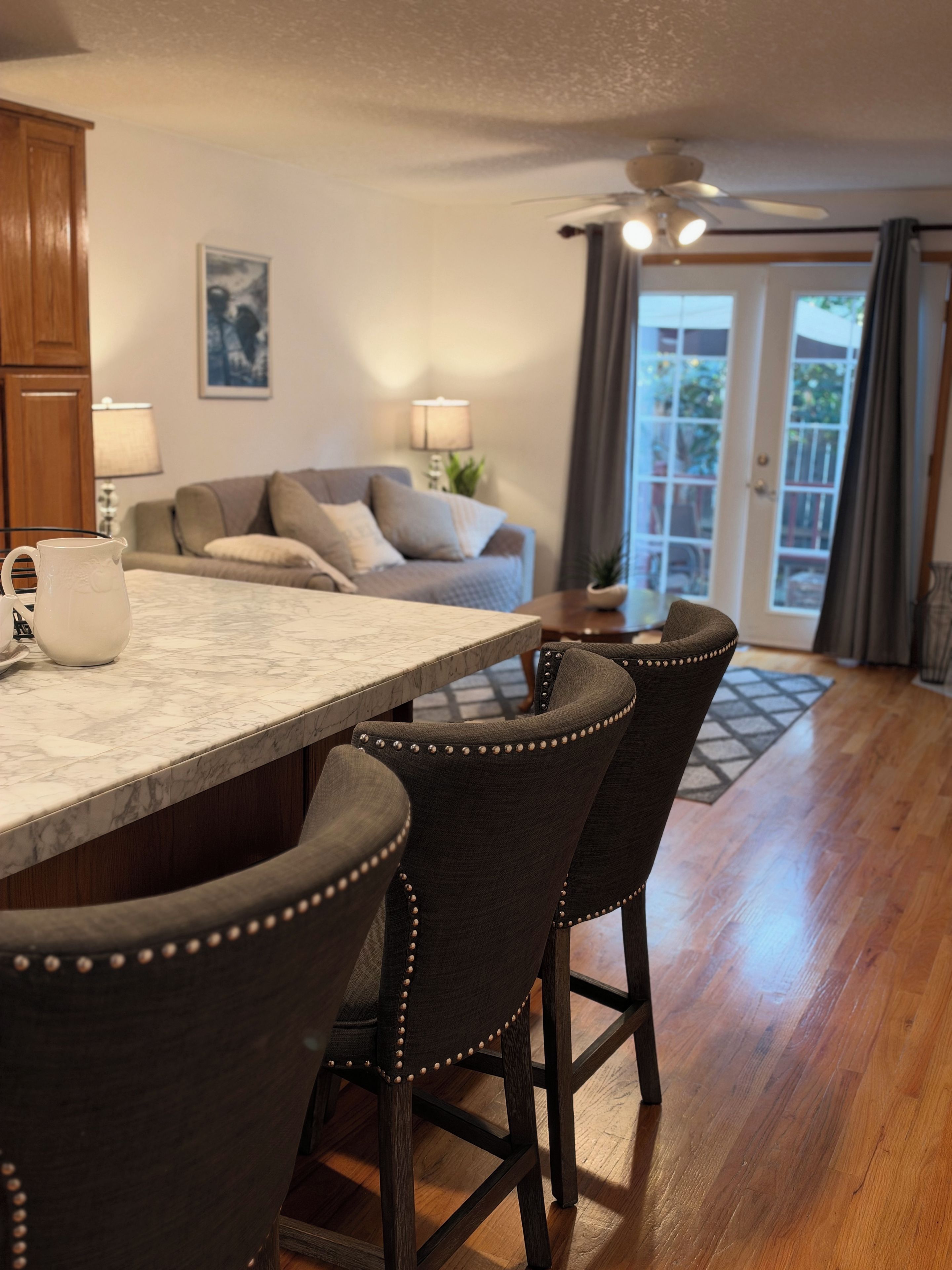 A kitchen with a marble countertop, dark upholstered bar stools, wooden cabinets, and a ceiling fan.