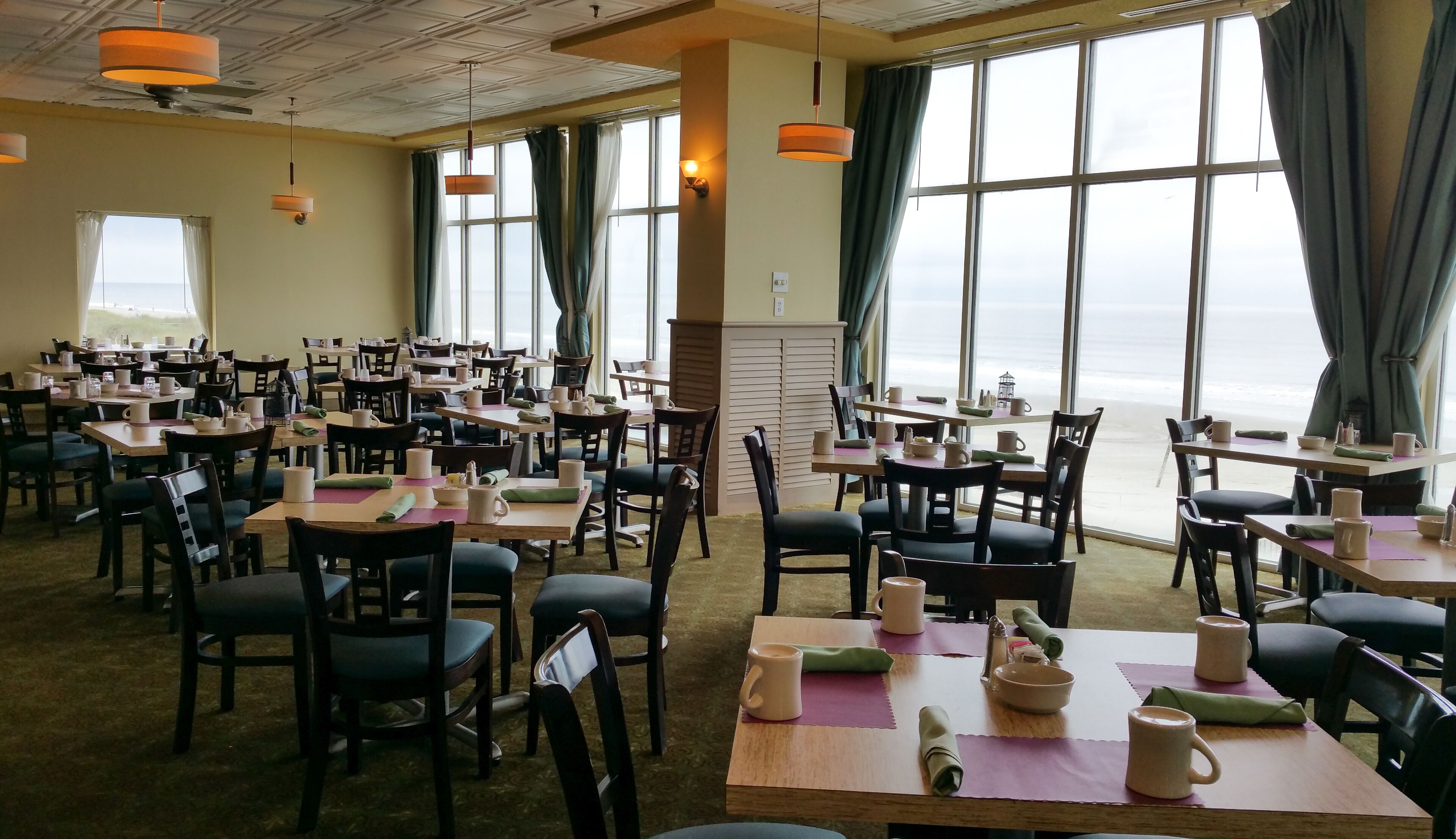 A dining area with tables set for service, large windows offering a view of the sea, and ceiling fans.