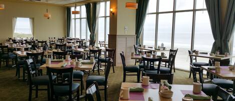 A dining area with tables set for service, large windows offering a view of the sea, and ceiling fans.