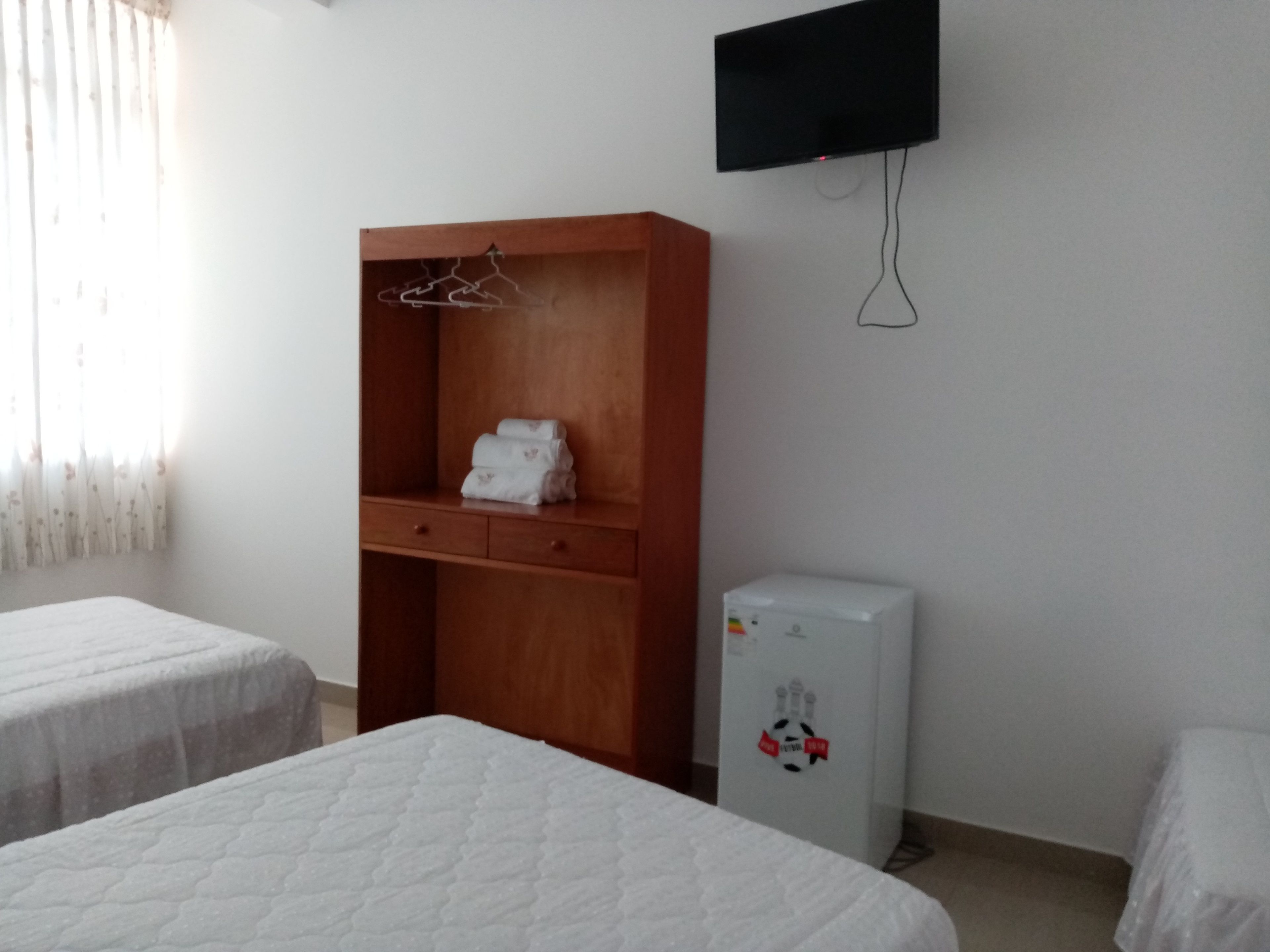Une chambre avec deux lits, une armoire en bois, un petit réfrigérateur et un téléviseur fixé au mur.