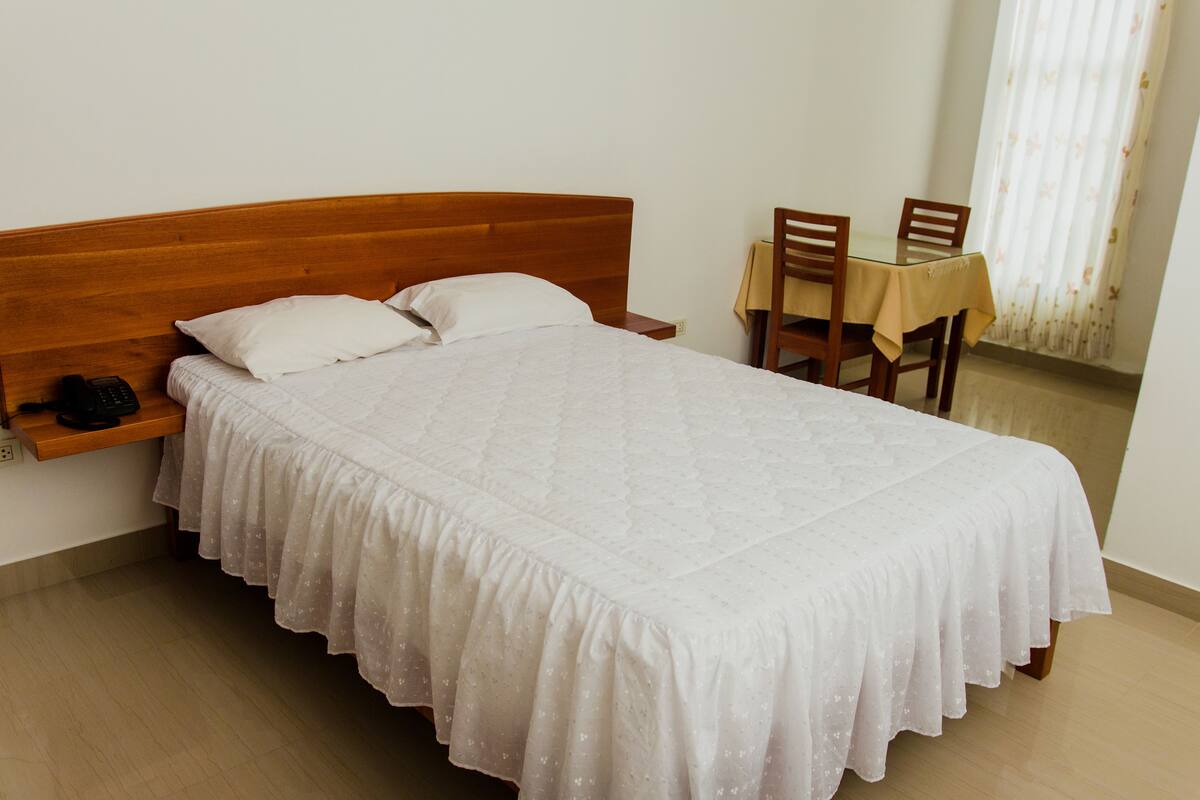 Cama bem arrumada com cabeceira de madeira, mesa de cabeceira com telefone e uma pequena prateleira. Há uma cadeira de madeira e uma mesa com toalha de mesa no canto.
