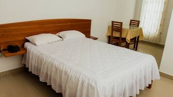 Cama bem arrumada com cabeceira de madeira, mesa de cabeceira com telefone e uma pequena prateleira. Há uma cadeira de madeira e uma mesa com toalha de mesa no canto.
