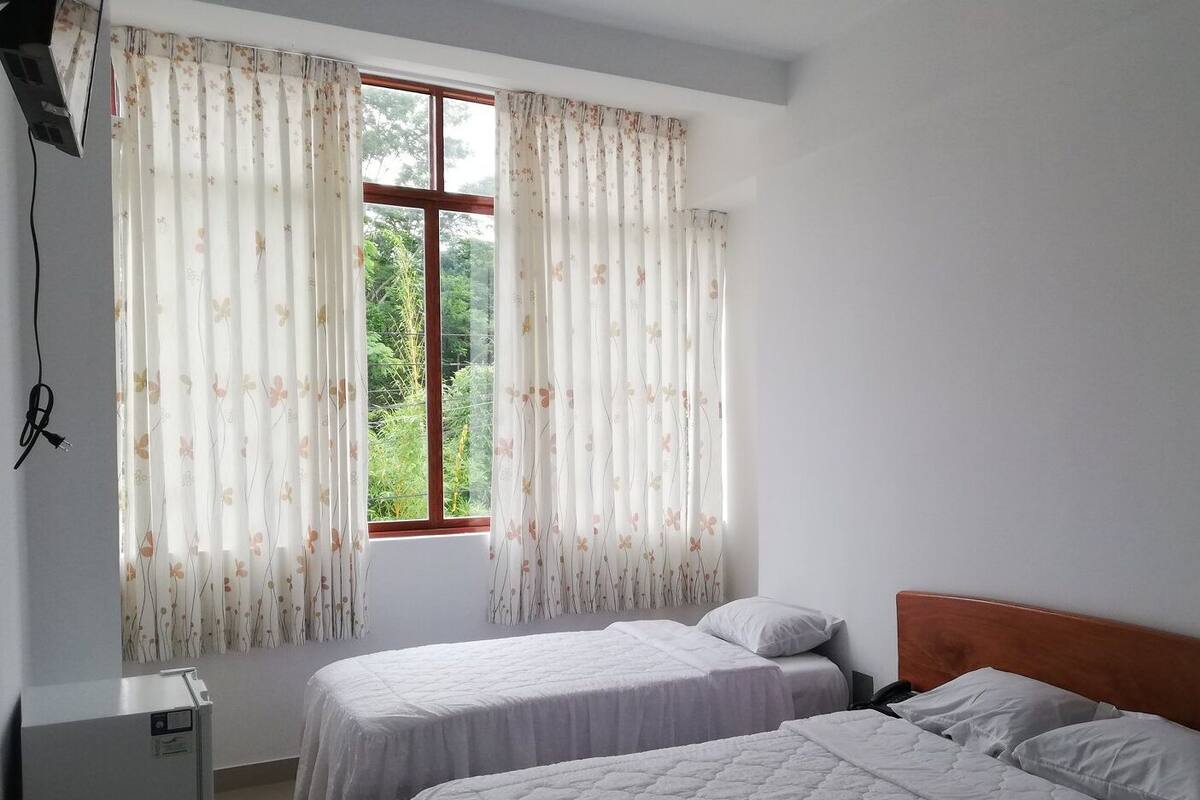 Quarto com duas camas, janela com cortinas florais e um refrigerador.
