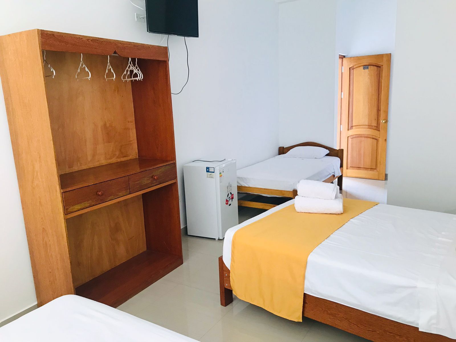 A room with a wooden wardrobe, a small refrigerator, two beds with white bedding, and a wooden door.